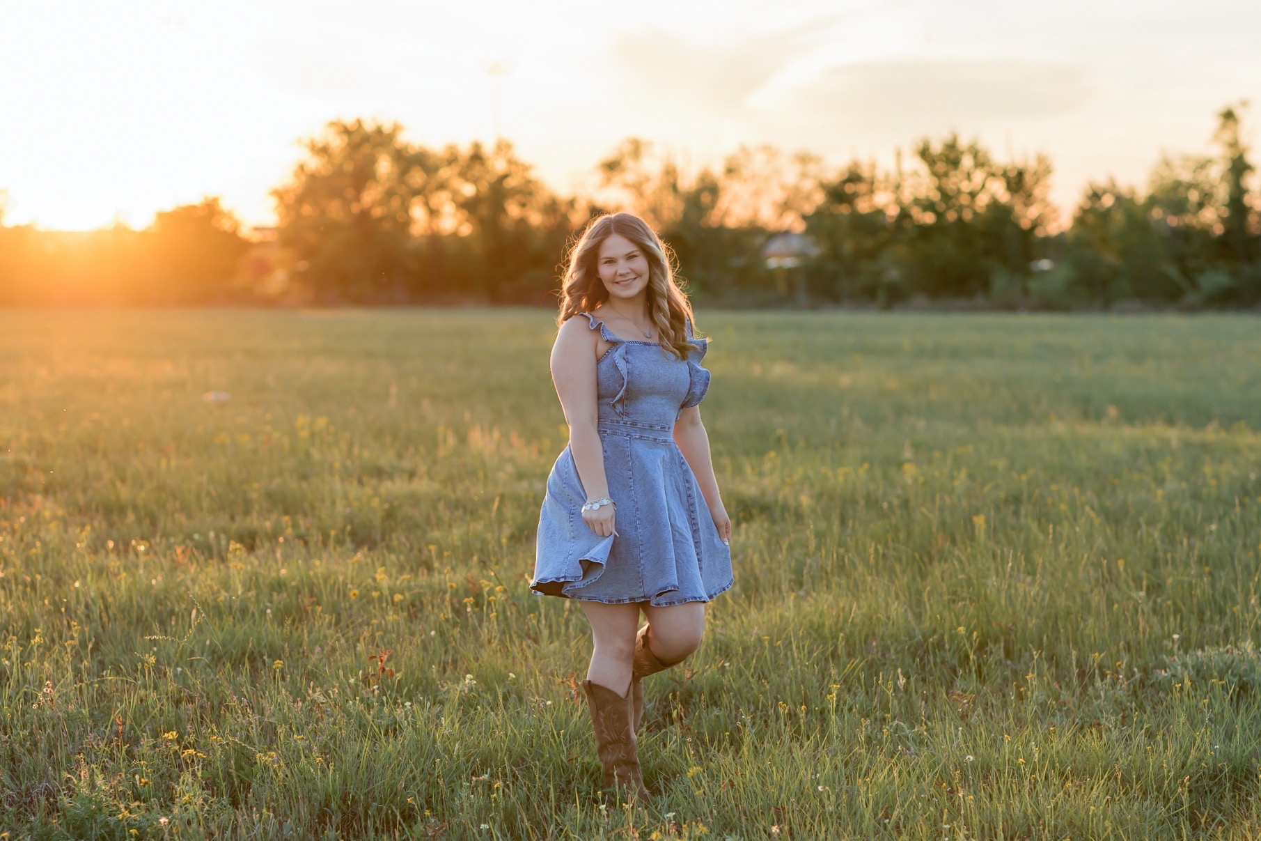 Senior in a denim dress and boots in a field at golden hour